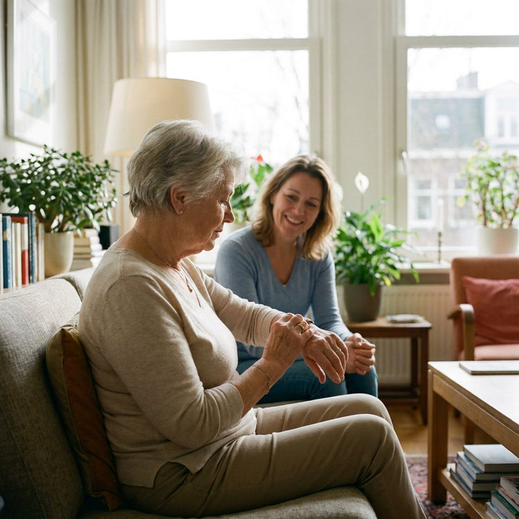 Oudere vrouw zit rustig in een lichte Nederlandse woonkamer en kijkt naar haar pols, terwijl een dochter op de achtergrond betrokken toekijkt.