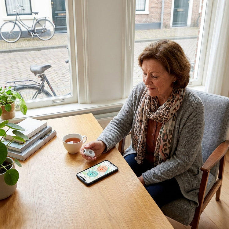 Oudere vrouw test een mobiele alarmknop bij de voordeur van haar woning, terwijl op tafel een smartphone met locatiekaart ligt in zacht daglicht.